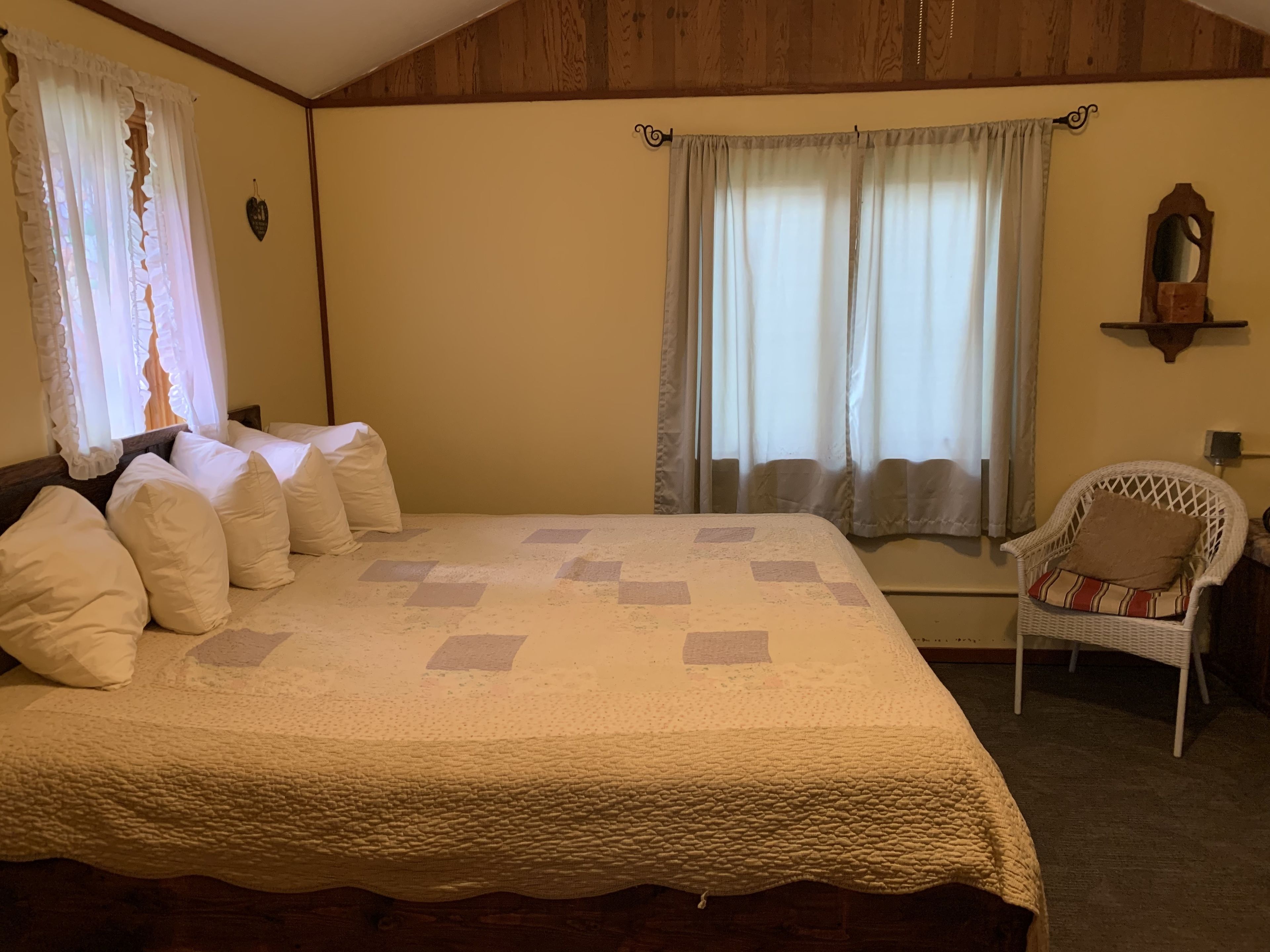 A bedroom with a large bed, two windows with curtains, a chair, and a wooden ceiling.