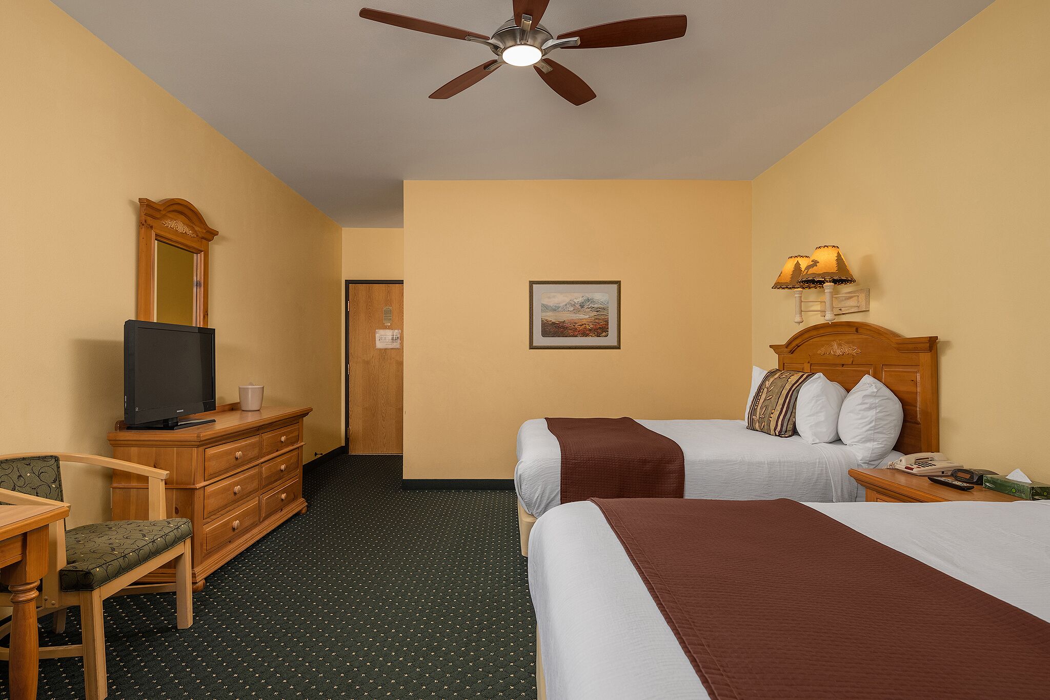 A hotel room with two beds, a television on a dresser, a desk, and a ceiling fan.