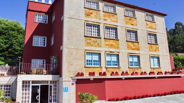 A multi-story building with a red facade and beige upper sections. It features multiple windows, a balcony with potted plants, and a small entrance area. - Hotel Xacobeo (Sanxenxo)