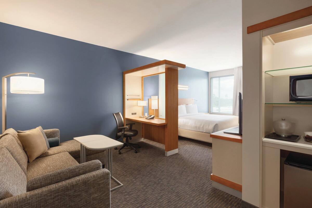 A hotel room with a sofa, a desk, a chair, a bed, a TV, and a small table.