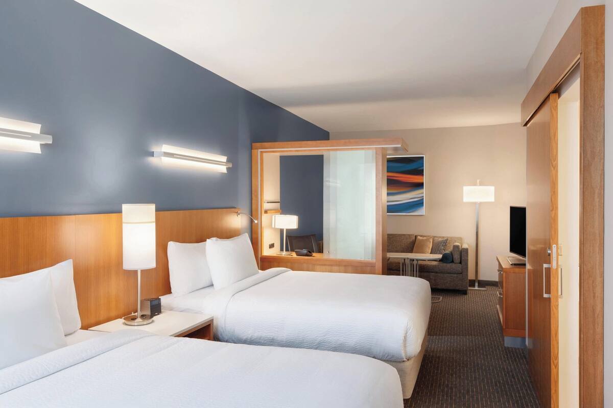 A hotel room with two beds, a wooden headboard, bedside lamps, a desk with a lamp, a television, a sofa, and a large mirror.