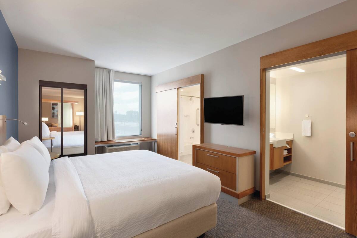 A hotel room with a large bed, a desk with a chair, a television mounted on the wall, a bathroom with a shower and toilet, and a sliding door closet.