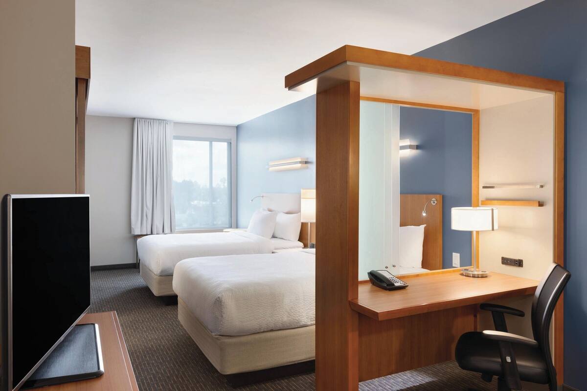 A hotel room with two beds, a desk with a chair, a TV, and a large mirror.