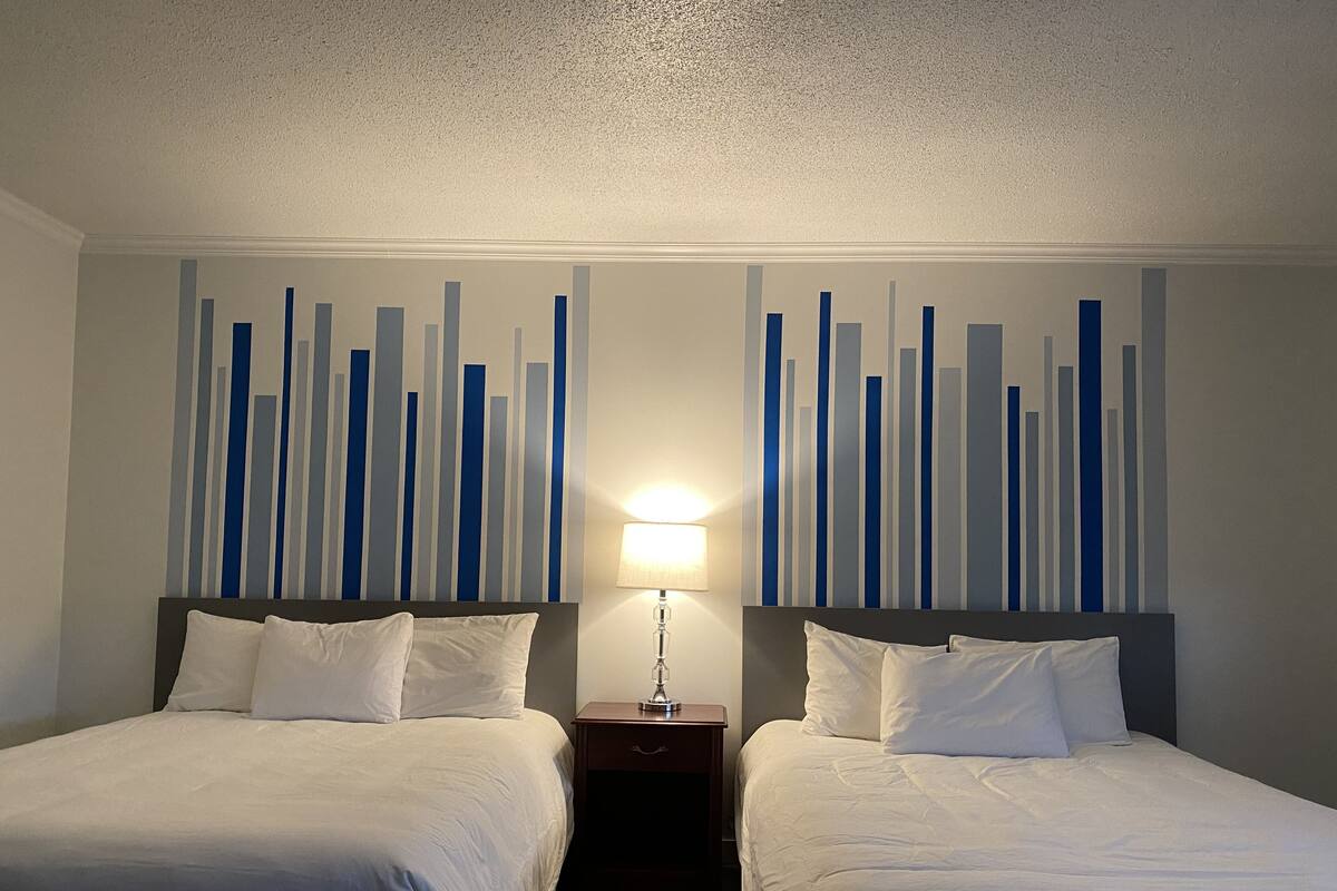 A hotel room with two beds, a nightstand with a lamp, and a wall with vertical blue and white stripes.
