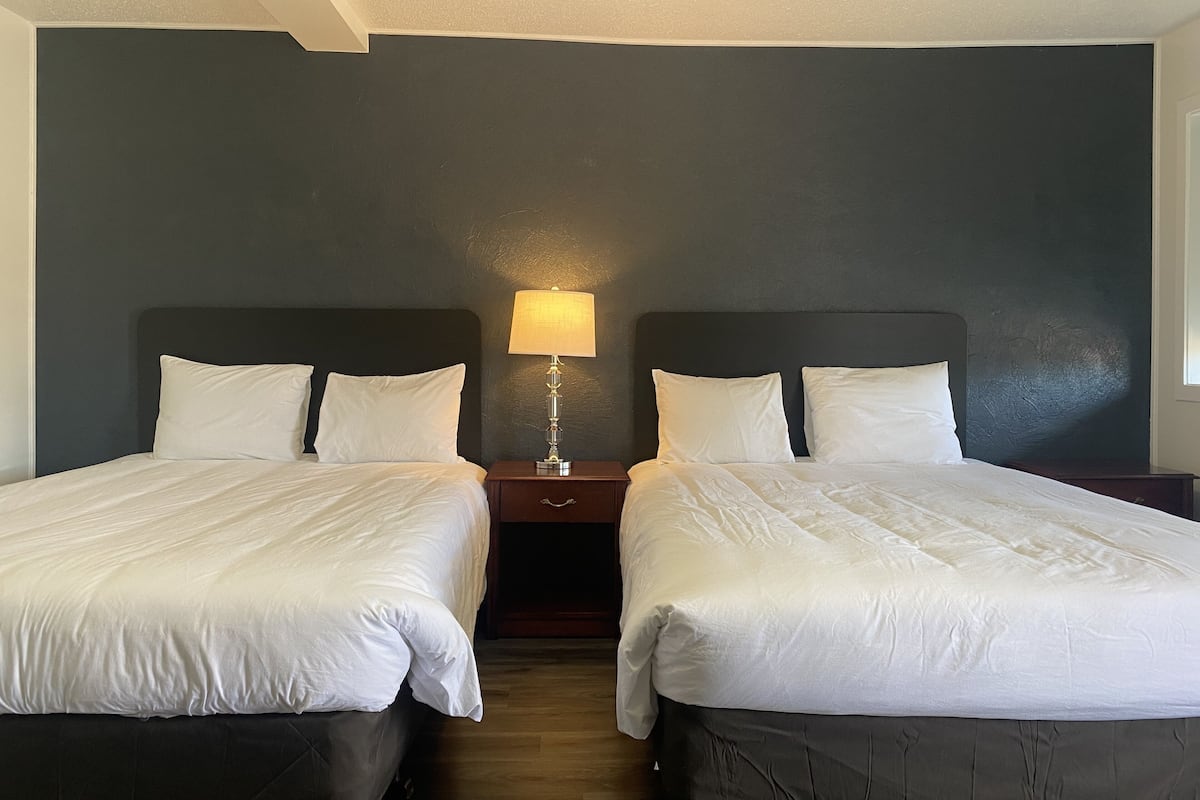 A hotel room with two beds, a nightstand with a lamp, and a dark-colored wall.