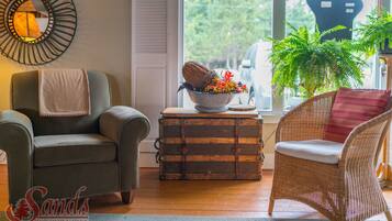 A living room with a grey armchair, a wicker chair with a red cushion, a wooden chest of drawers, a mirror, and a potted plant.