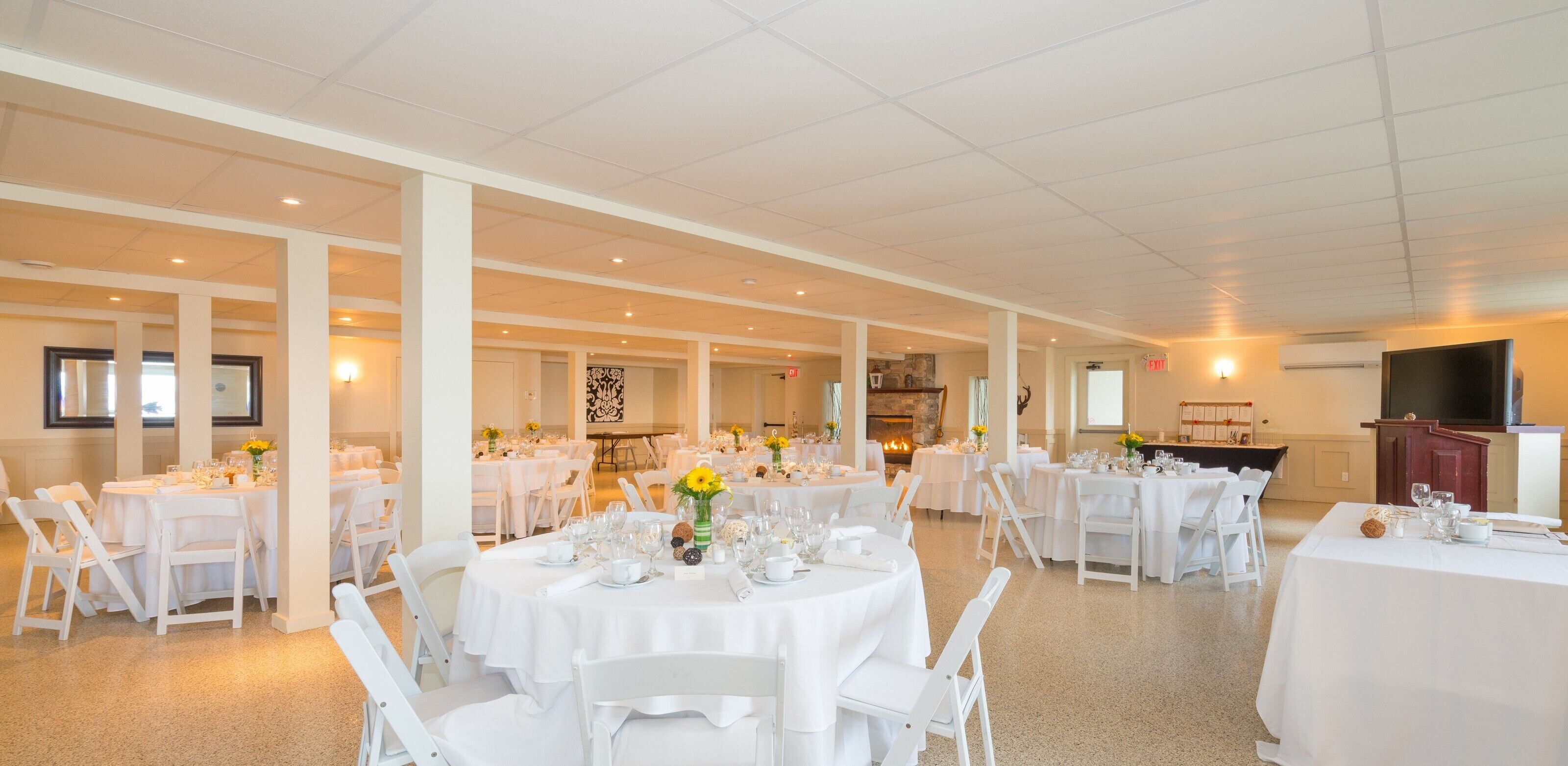 A spacious banquet hall with multiple round tables set for dining, white chairs, and a large flat-screen TV mounted on the wall.