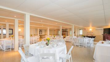A spacious banquet hall with multiple round tables set for dining, white chairs, and a large flat-screen TV mounted on the wall.