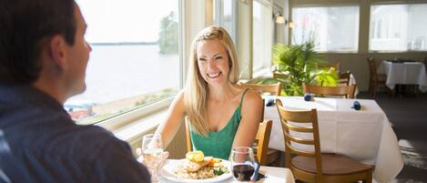 A dining area with a table set for two, a view of the water, and a potted plant.