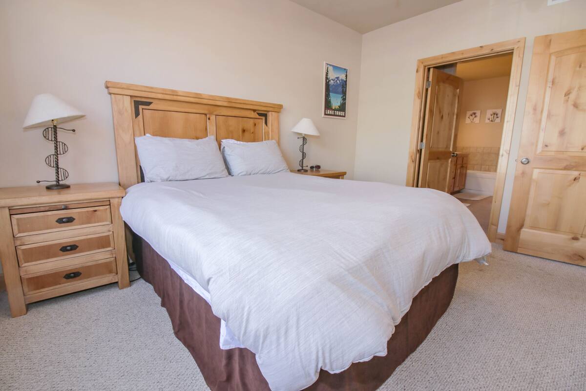 A bedroom with a large bed, wooden headboard, two bedside tables with lamps, a wooden door, and a bathroom visible through an open door.