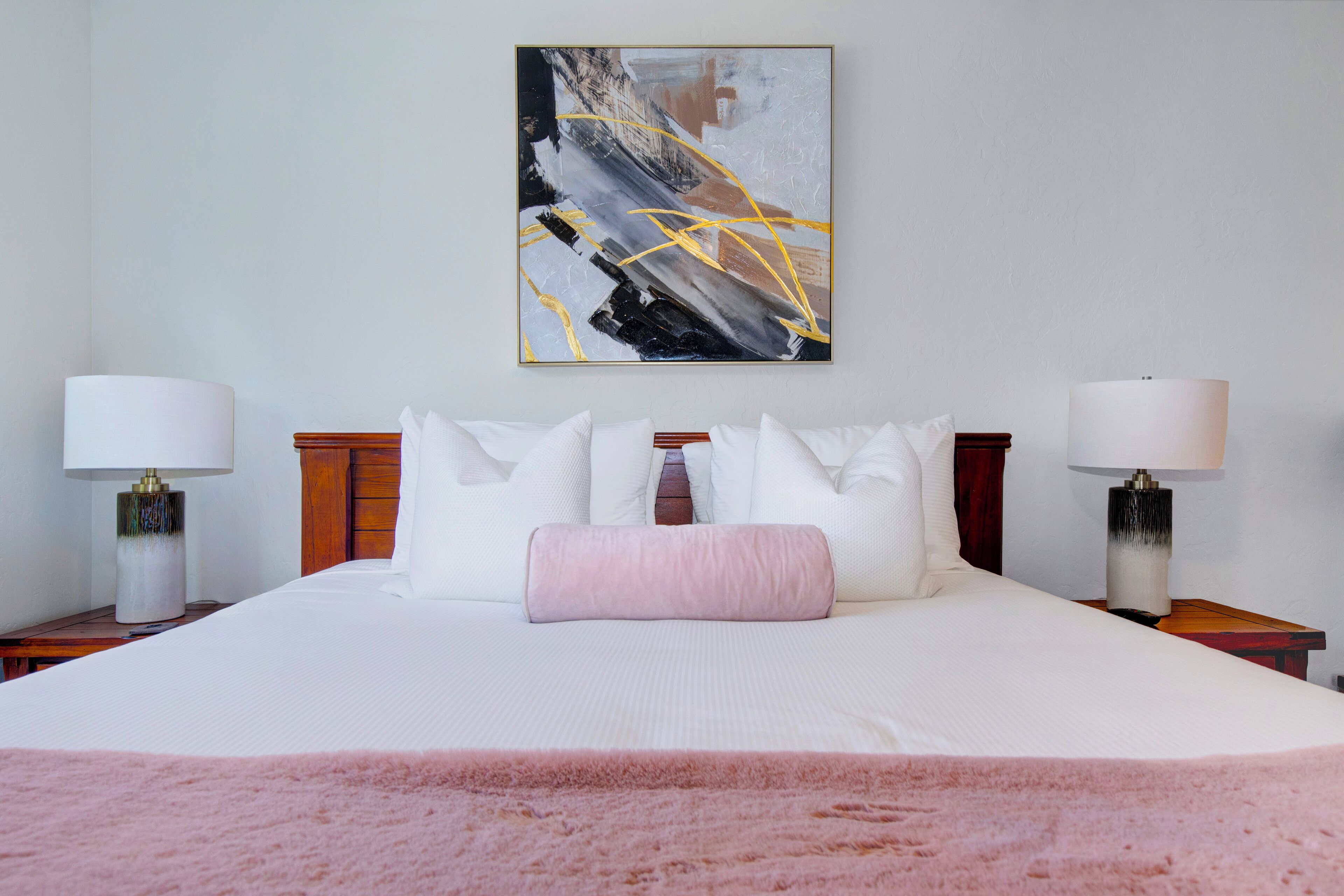 Ein Hotelzimmer mit einem großen Bett, weißer Bettwäsche, einer rosafarbenen Tagesdecke, zwei Nachttischen mit Lampen und einem modernen abstrakten Gemälde an der Wand.