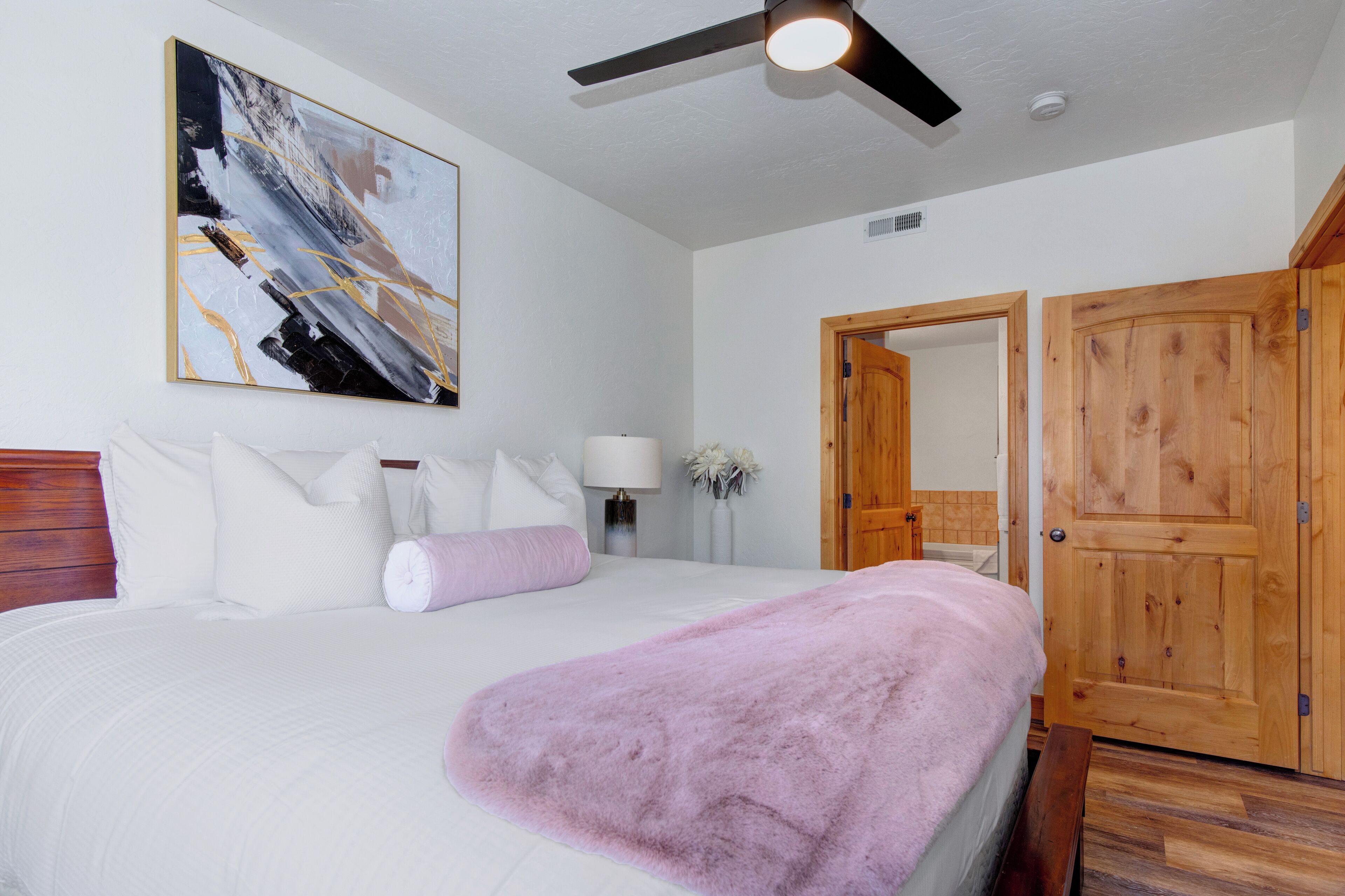 A bedroom with a bed, a ceiling fan, a wooden door, and a painting on the wall.