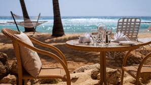 Montaje para comer en la playa con sillas de mimbre y una mesa para dos, con vista al océano.