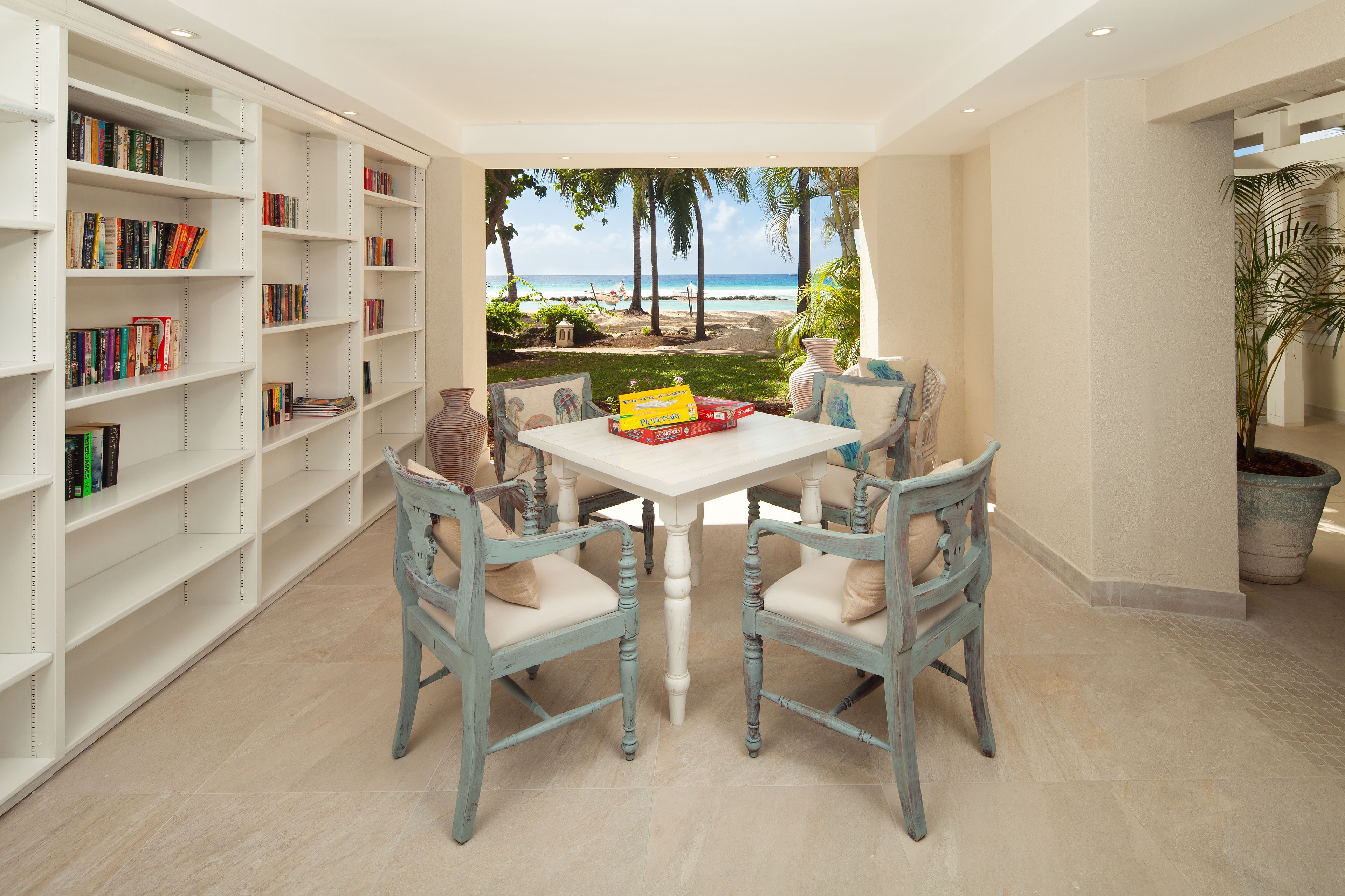 A room with a view of the beach, a white table with chairs, and a bookshelf filled with books.