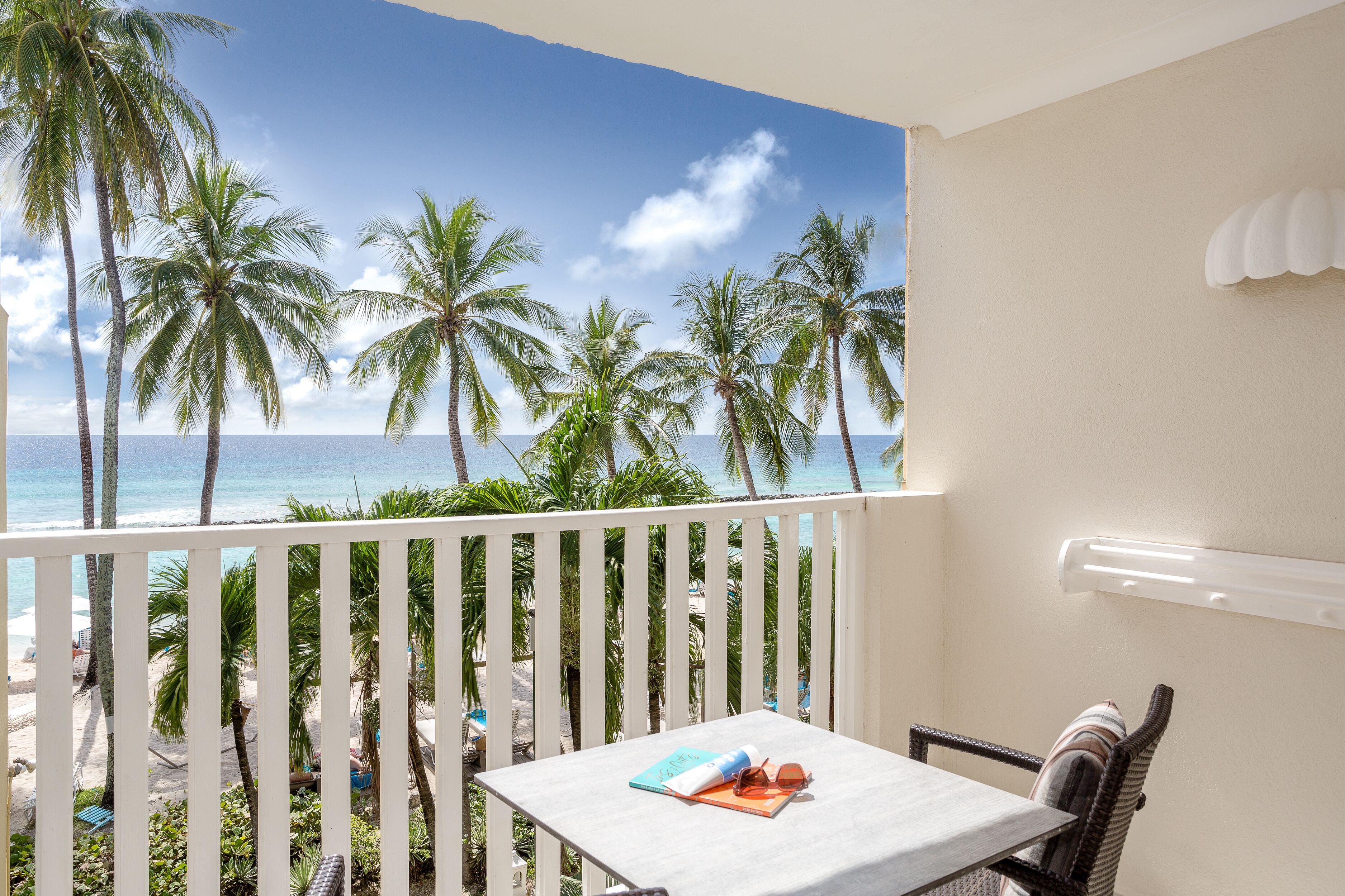 A balcony with a view of the beach and palm trees, a table with a book and sunglasses, and a white railing.