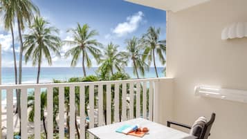 A balcony with a view of the beach and palm trees, a table with a book and sunglasses, and a white railing.