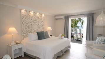 Una habitación de hotel con una cama grande, dos mesitas de noche, una silla, un balcón con mesa y sillas, y una lámpara colgante.