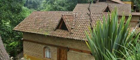 A rustic brick house with a tiled roof, surrounded by lush greenery and a tree.