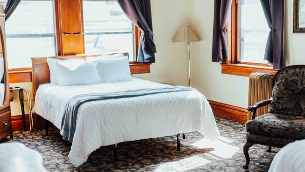 A hotel room with a large bed, dark curtains, a wooden floor, and a chair. - Town Hall Inn (Lead)
