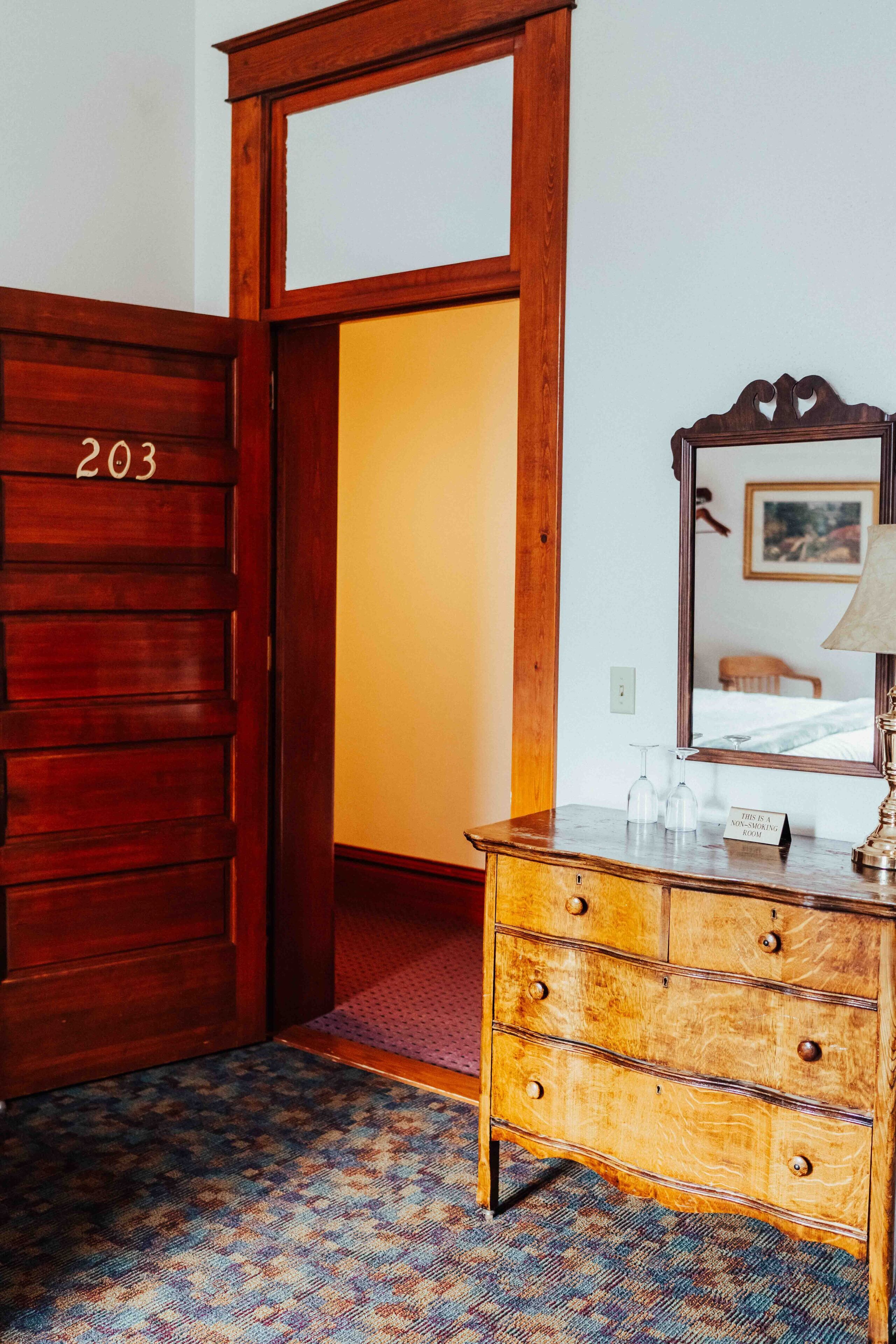 Une porte en bois portant le numéro 203, une commode en bois avec des tiroirs, un miroir et un panneau indiquant que la chambre n’est pas disponible.