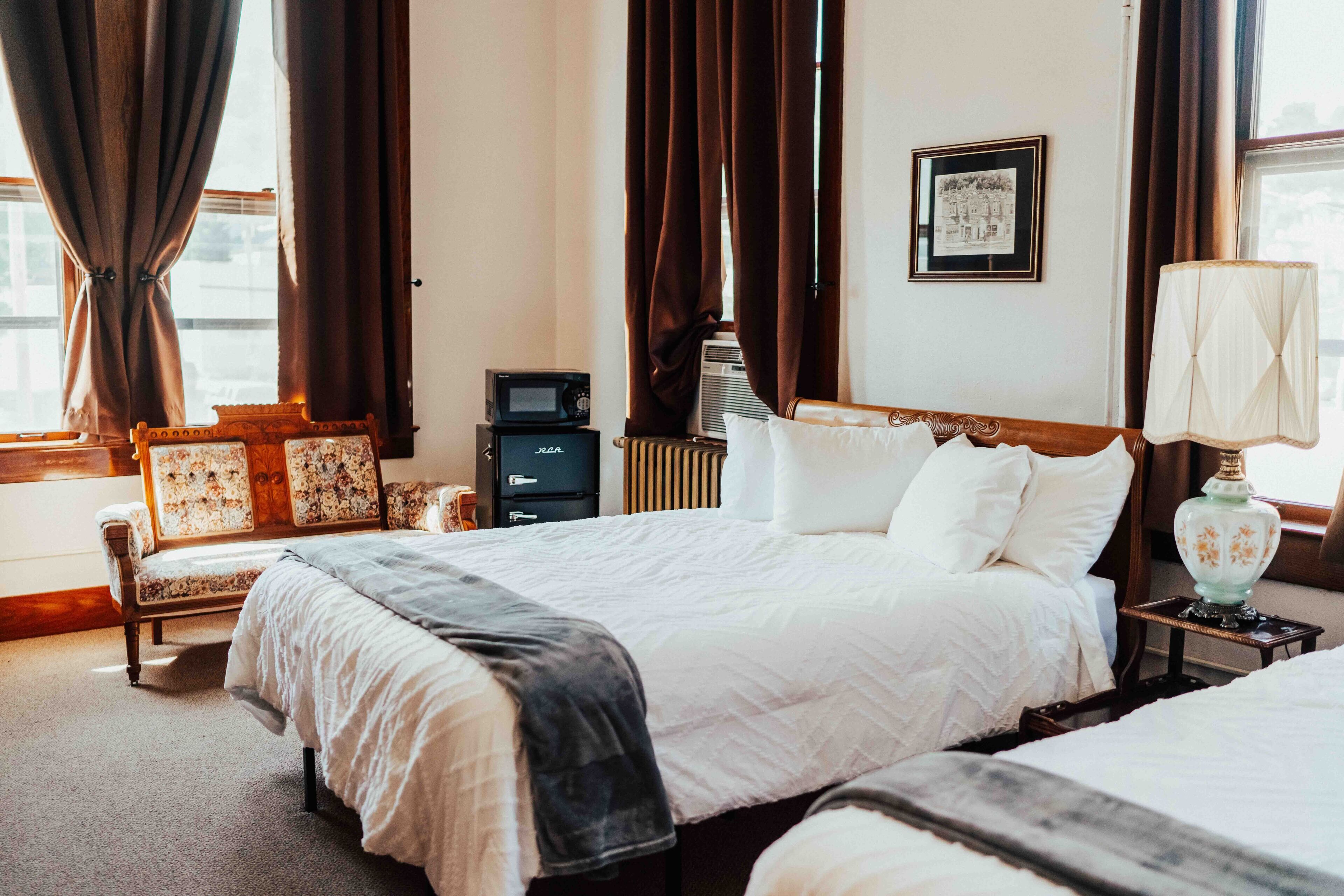 A hotel room with two beds, a desk, a chair, a radiator, a framed picture on the wall, and a window with curtains.