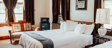 A hotel room with two beds, a desk, a chair, a radiator, a framed picture on the wall, and a window with curtains.