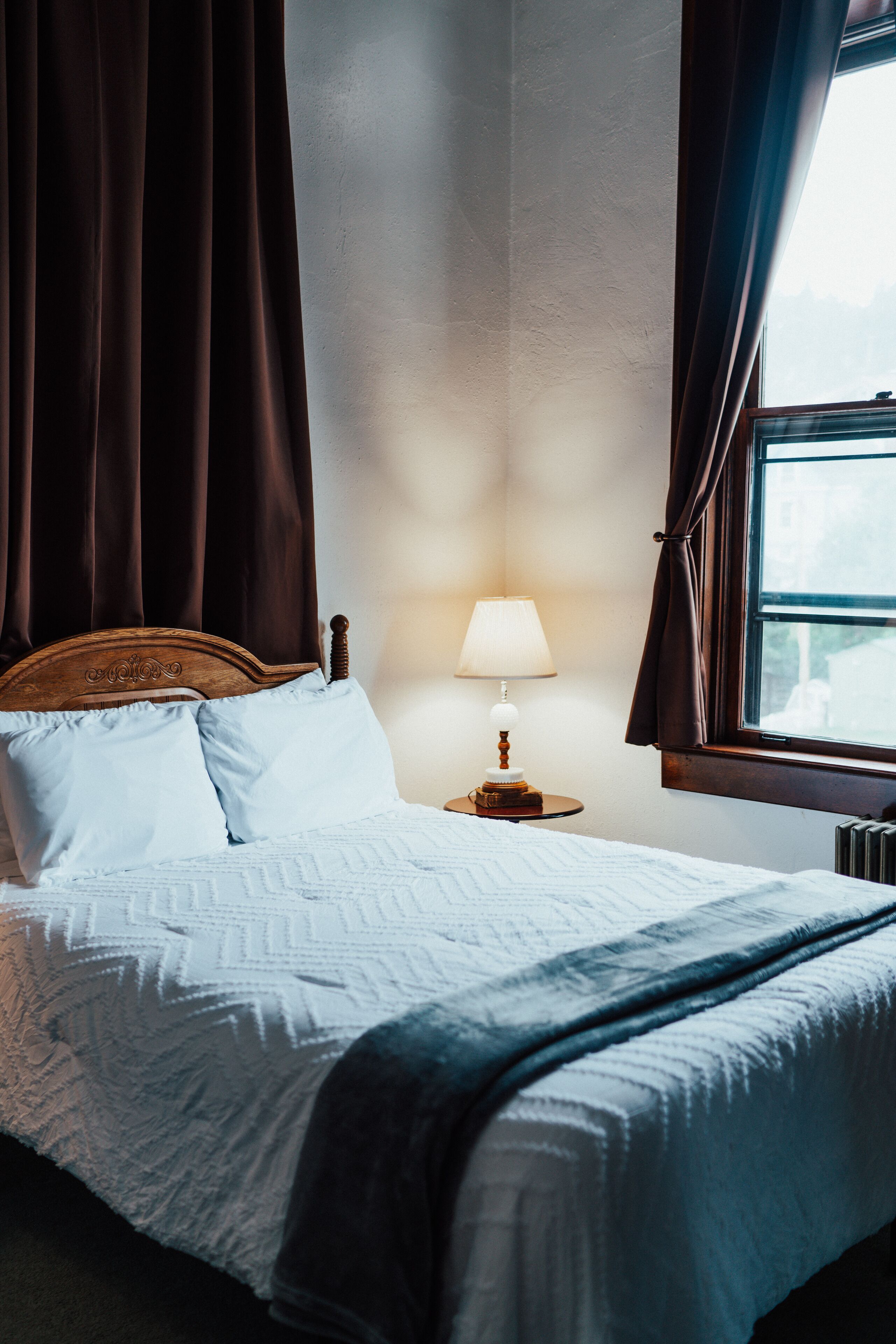 Una cama bien hecha con cabecera de madera, una lámpara de noche y una ventana con cortinas.