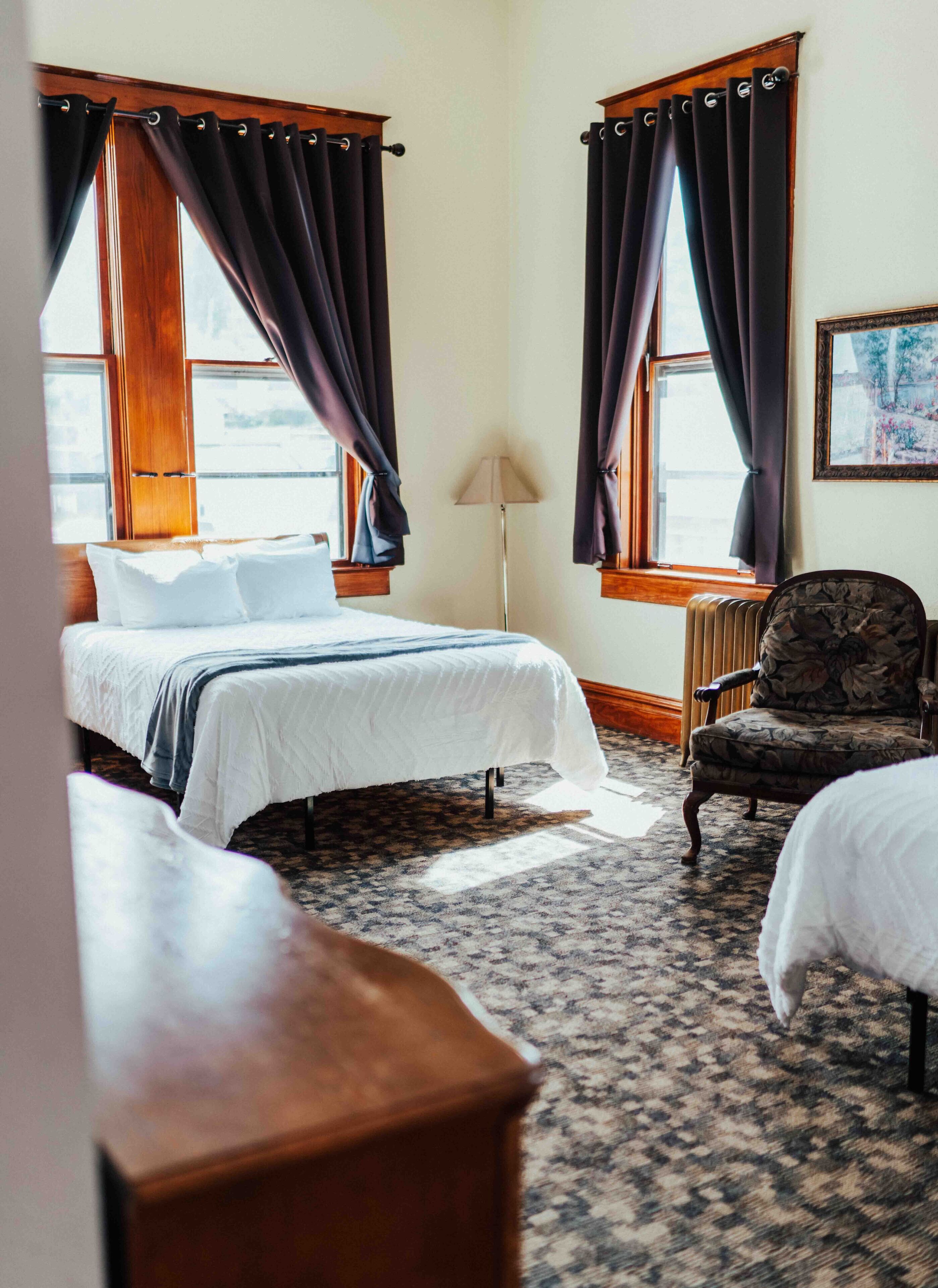 Een hotelkamer met een groot bed, twee ramen met donkere gordijnen, een stoel, een lamp en een kamerbreed tapijt met dessin.