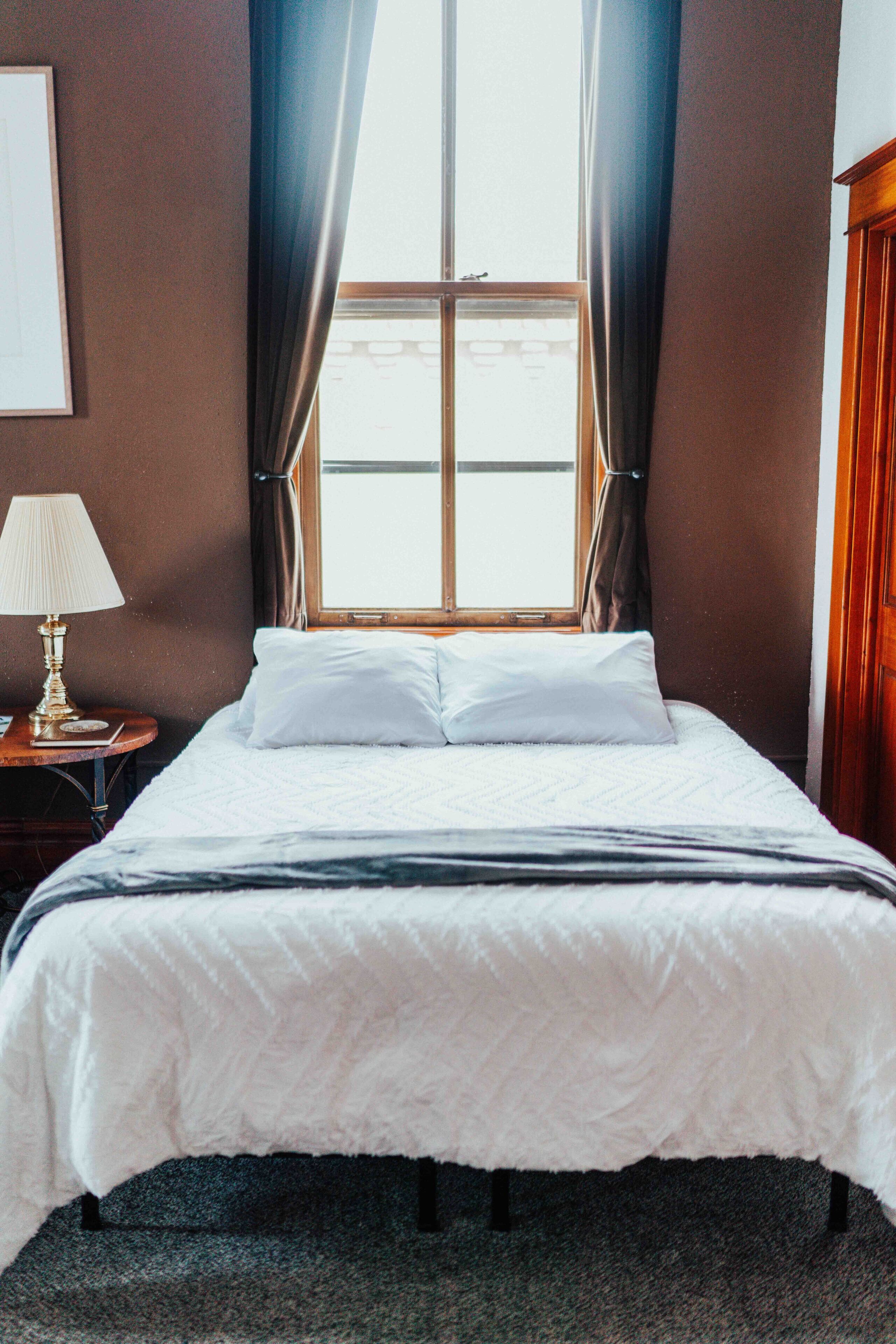 A neatly made bed with white linens, a bedside lamp, and a window with curtains.