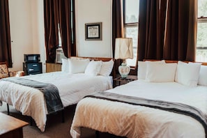 A hotel room with two beds, a bedside table, a lamp, a radiator, and a framed picture on the wall.