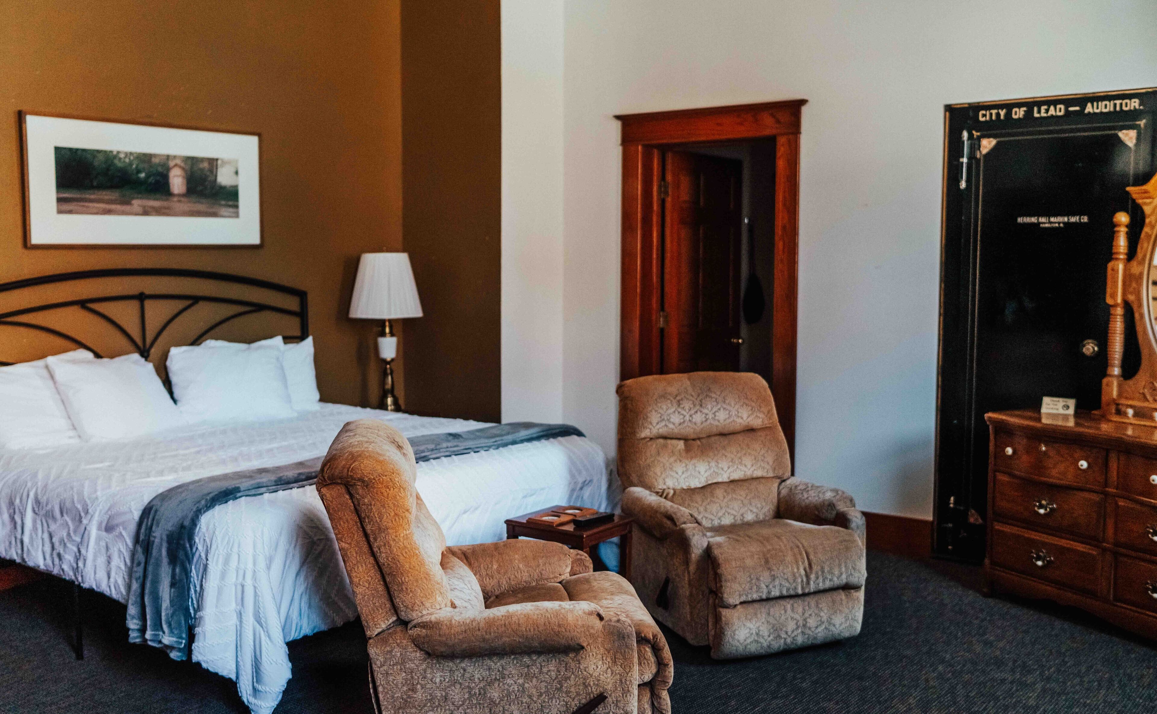 Habitación de hotel con cama, dos sillones, mesita de noche, lámpara, cómoda y puerta.
