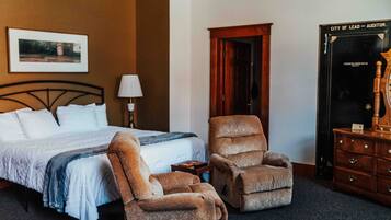 Habitación de hotel con cama, dos sillones, mesita de noche, lámpara, cómoda y puerta.