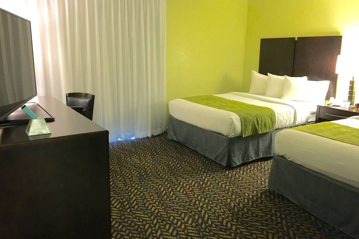Quarto de hotel com cama, escrivaninha, cadeira e uma janela grande com cortinas.