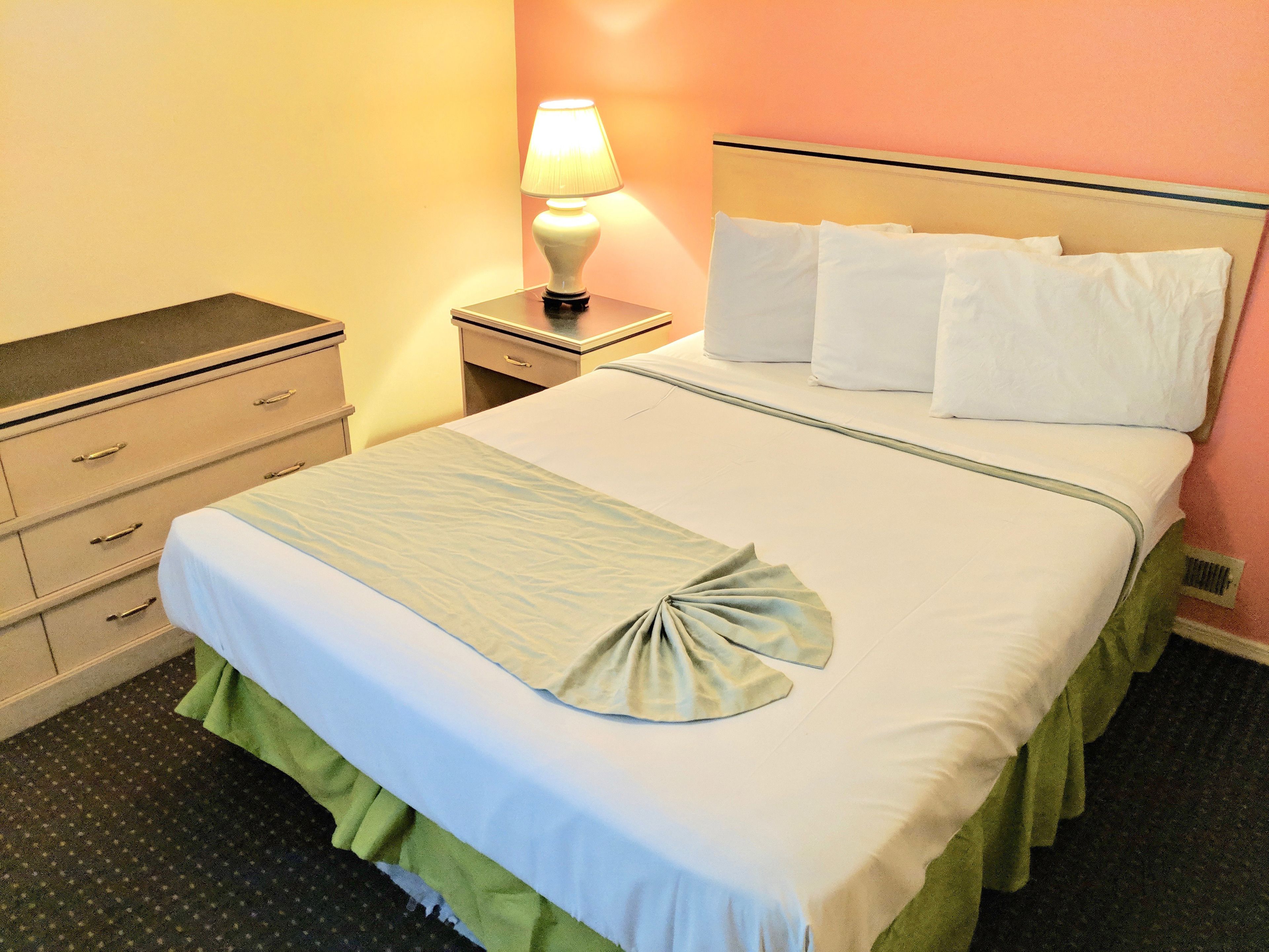 A neatly made bed with white linens and a green skirt, a bedside lamp, and a nightstand with a drawer.