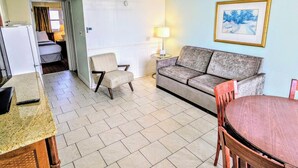 A compact hotel room with a kitchenette, a sofa, a chair, a dining table, and a painting on the wall. - Quebec Motel (Wildwood)