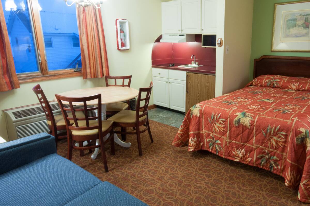 Une chambre d’hôtel compacte comprenant un lit, une petite kitchenette, une table à manger avec des chaises et une fenêtre avec des rideaux.