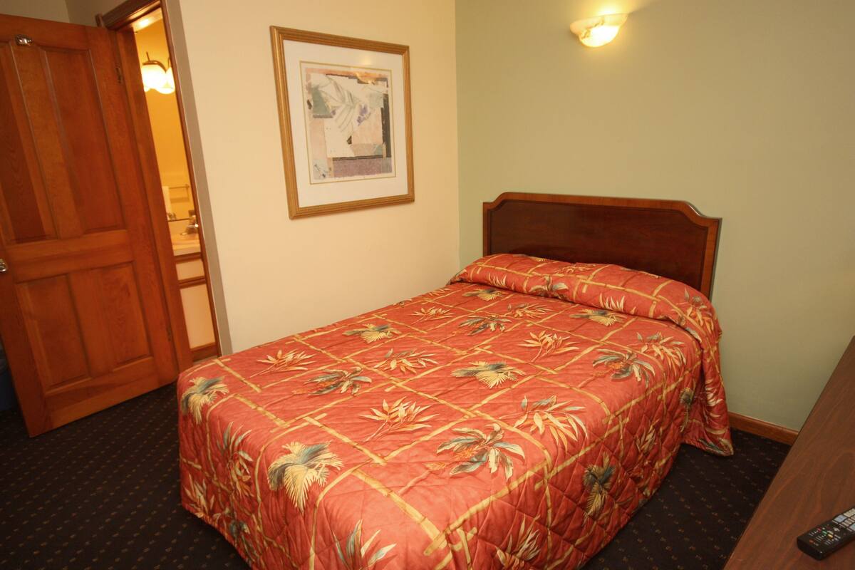 Une chambre d’hôtel avec un lit, une porte en bois, un tableau encadré au mur et une télécommande sur la table de chevet.