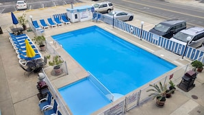 A rectangular pool with blue water, surrounded by a white and blue fence. There are lounge chairs and umbrellas along the poolside.
