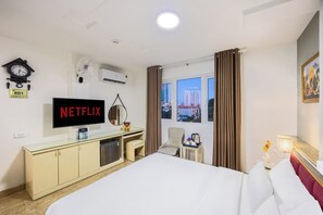 A hotel room with a bed, a desk with a computer, a television displaying 'NETFLIX', a clock, an air conditioning unit, a window with curtains, and a city view. - A25 Hotel - 22 Nguyen Cu Trinh (Ho Chi Minh City)