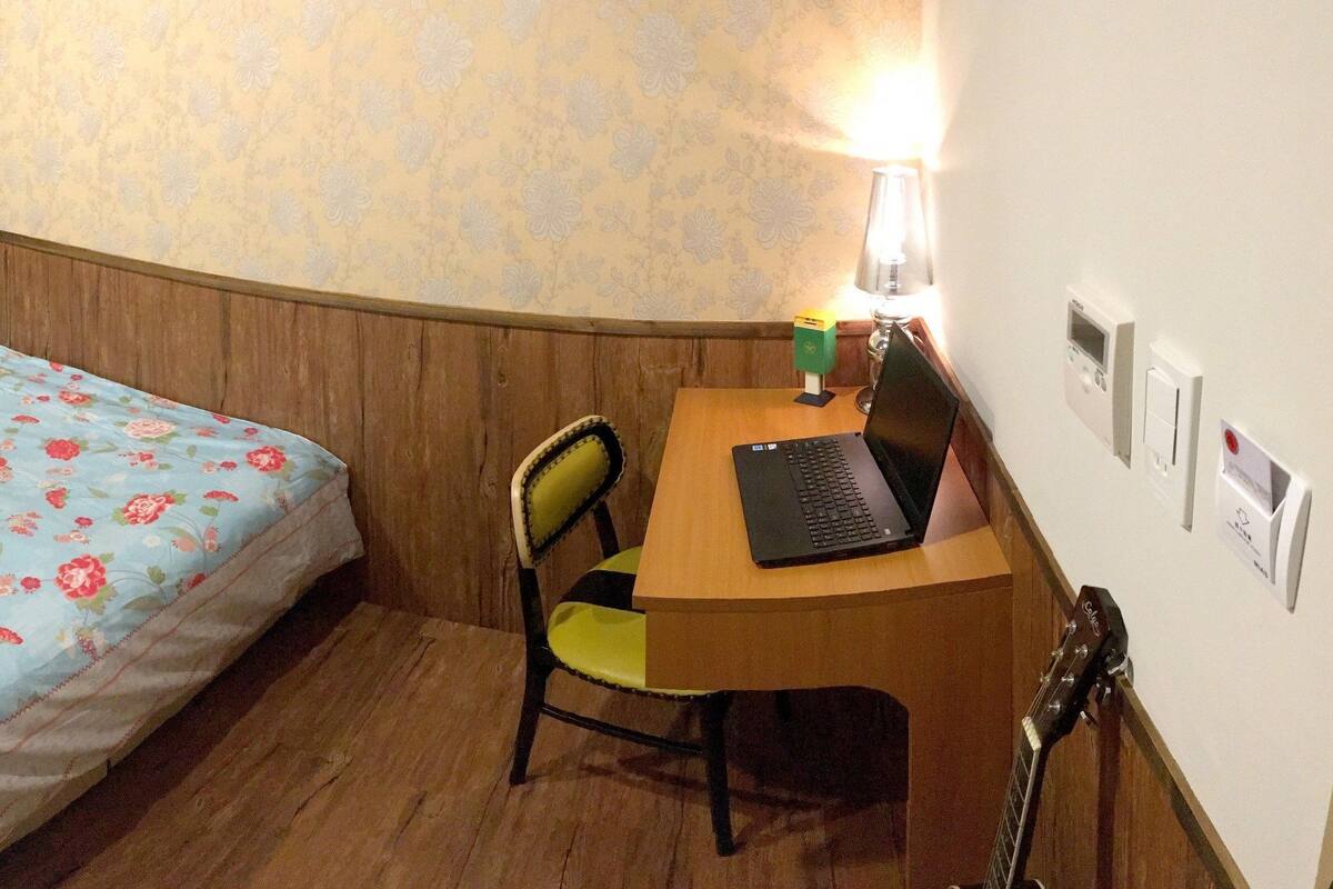 A bedroom with a bed, a desk with a laptop, a chair, a lamp, and a guitar.
