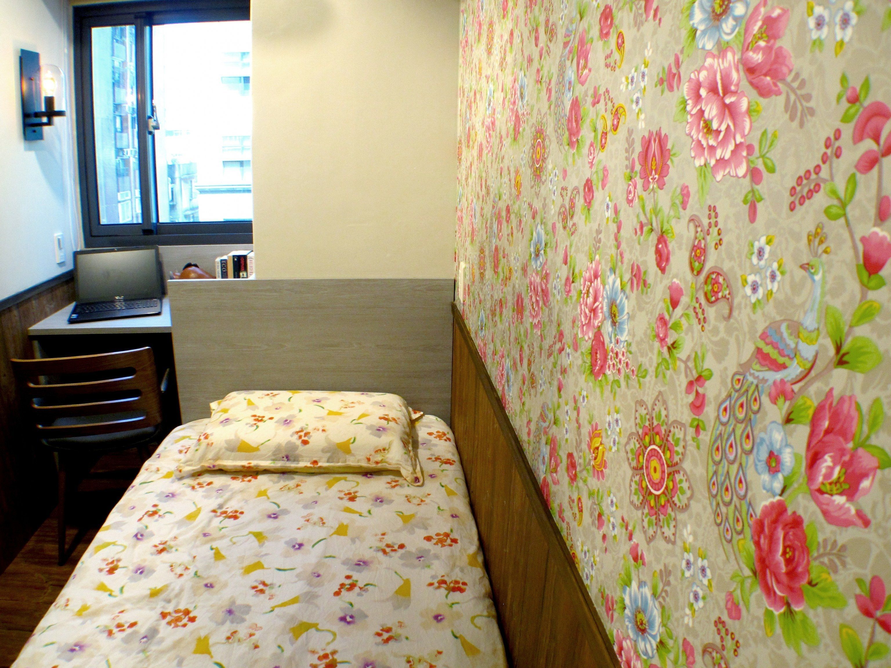 A single bed with floral bedding, a desk with a laptop, and a window with a view of buildings.