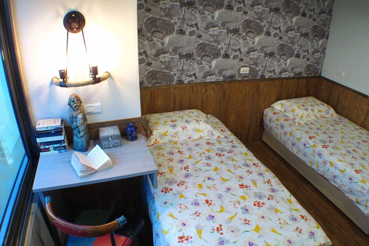 A room with two beds, a desk with books and a lamp, a stone wall, and a small window.