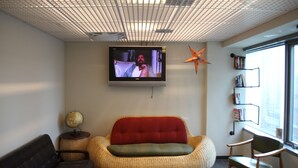 Ein Zimmer mit an der Wand montiertem Fernseher, einem Korbssessel, einem dunklen Ledersofa, einem Globus auf einem Beistelltisch und einem Bücherregal am Fenster.