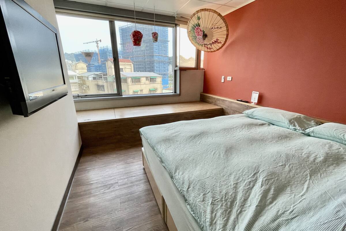 A bedroom with a bed, a window, a fan, and a TV mounted on the wall.