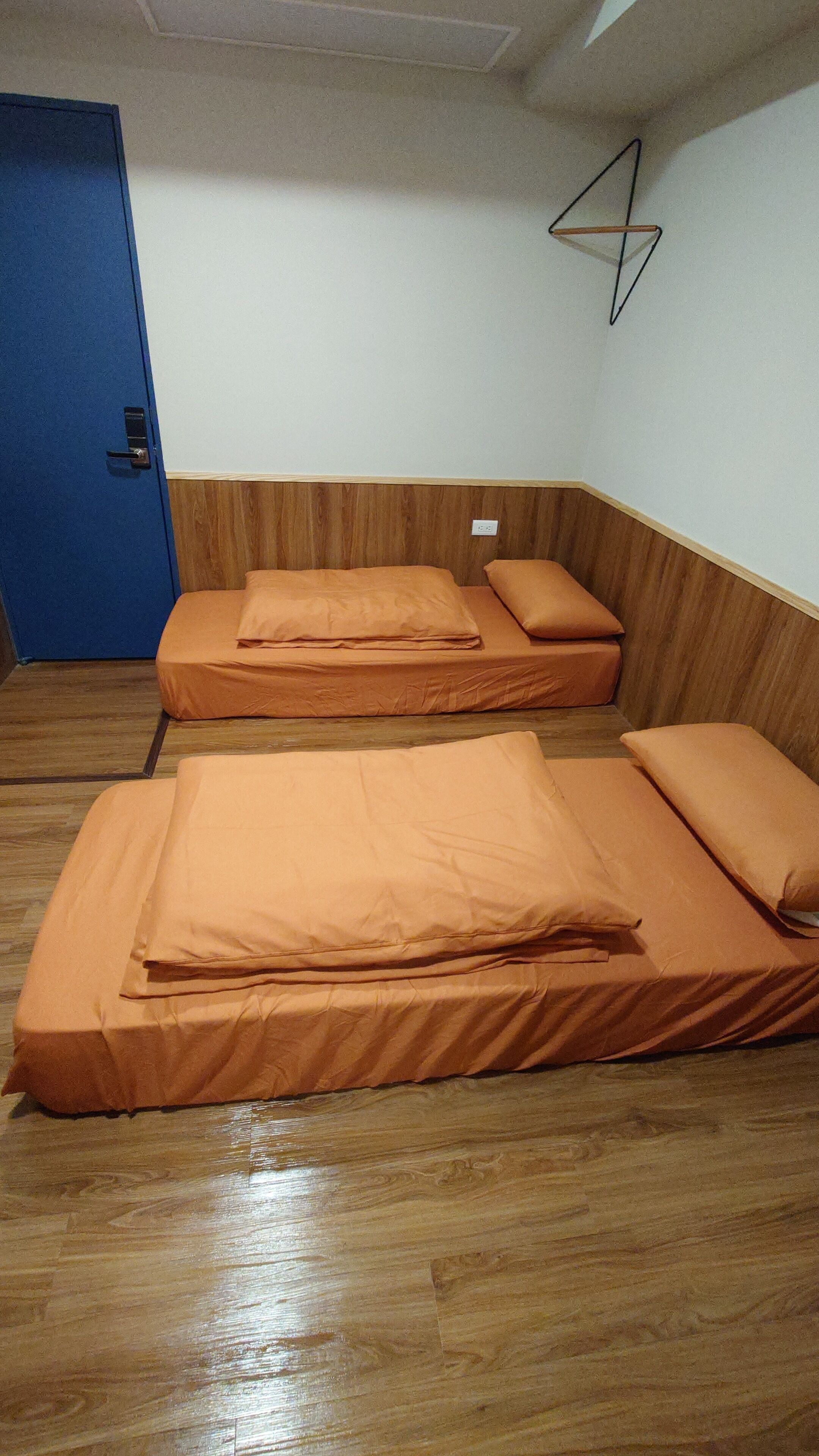 Una habitación con dos camas, suelo de madera, una puerta azul y un enchufe de pared.