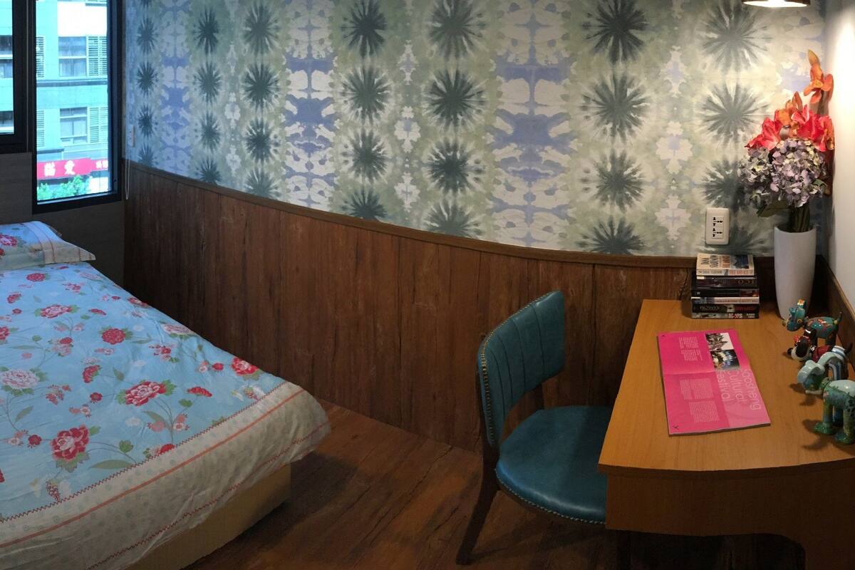 A bedroom with a bed, a desk, a chair, a vase with flowers, and a window with a view of buildings.
