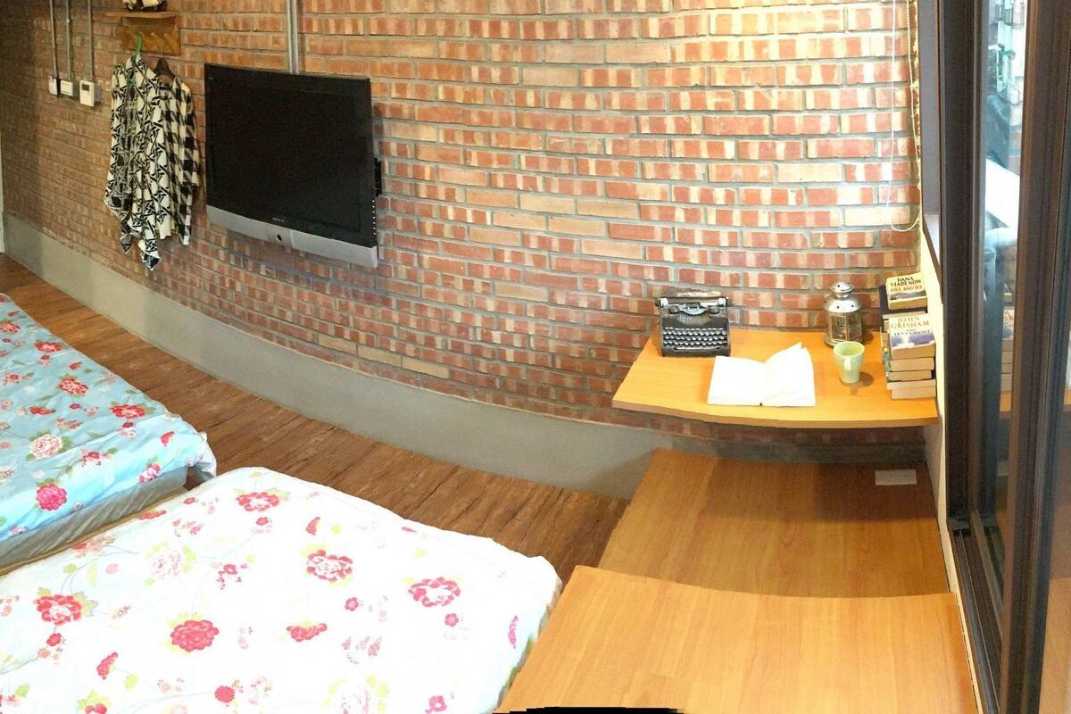 A small room with two beds, a brick wall, a flat-screen TV, and a small table with books and a lamp.