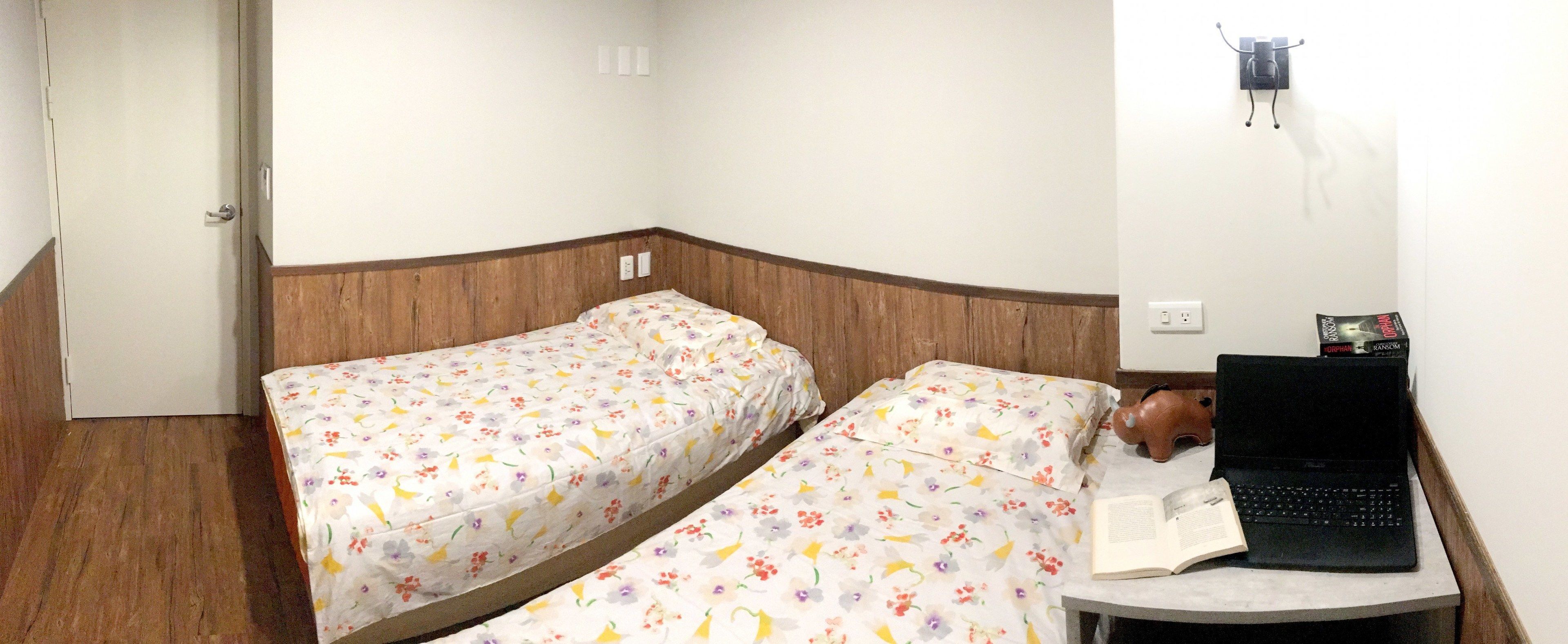A room with two beds, a small table with a laptop and a book, and a wall-mounted light fixture.
