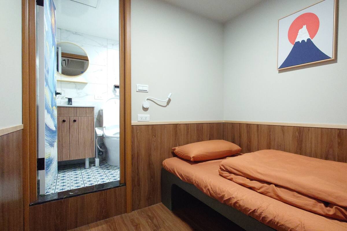 A bedroom with a bed, a wooden headboard, a wall-mounted light switch, and a framed picture of a mountain.