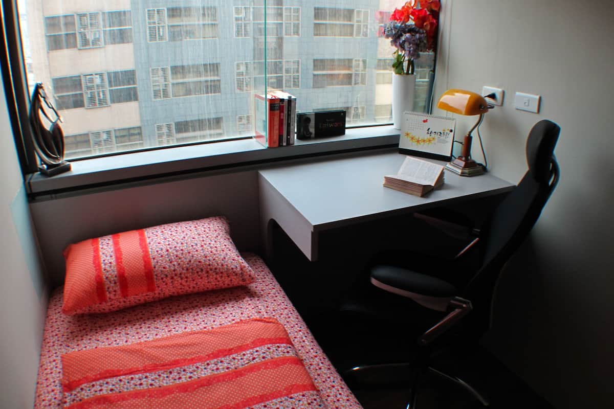 A small bedroom with a single bed, a desk, and a chair. There is a window with a view of buildings outside.